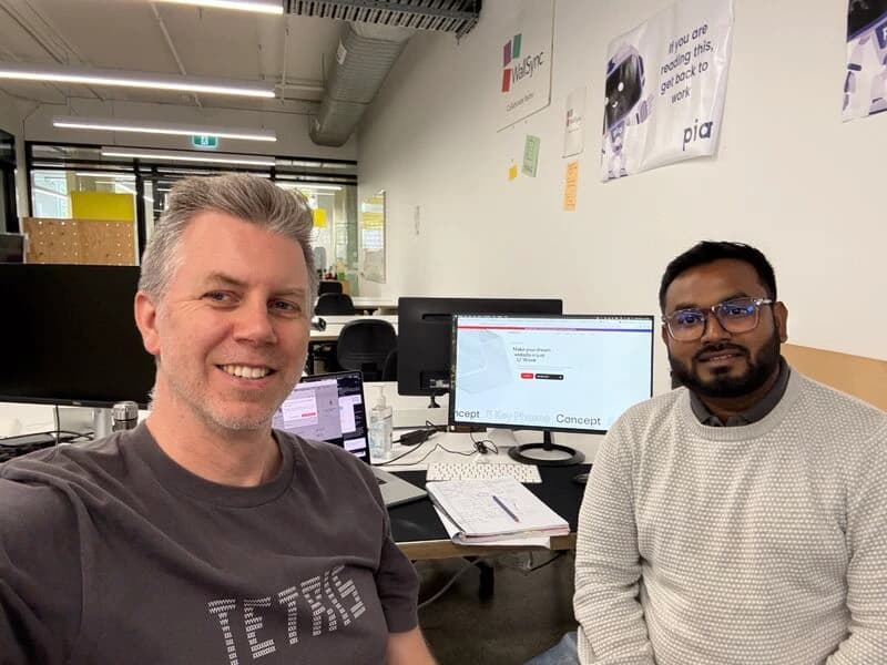 Two people at a desk in an open-plan office, a website design on the monitor behind them